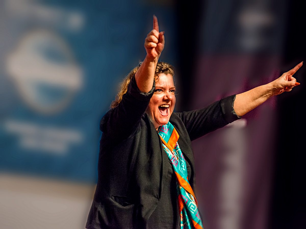 Jo-Dee Walmsley mid-keynote on stage, arms raised, pointing energetically out to the audience.