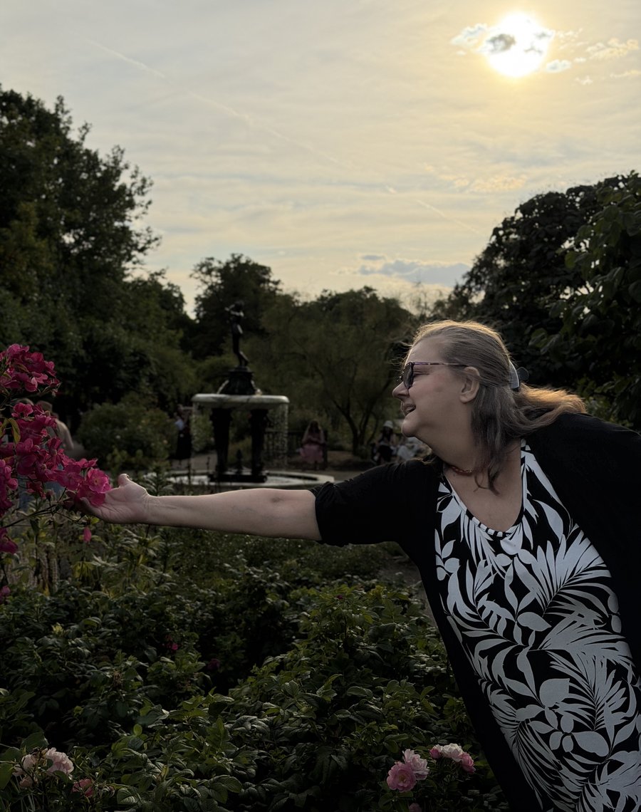 Jo-Dee Walmsley reaching out to touch pink roses in a garden, smiling, golden evening light behind her.