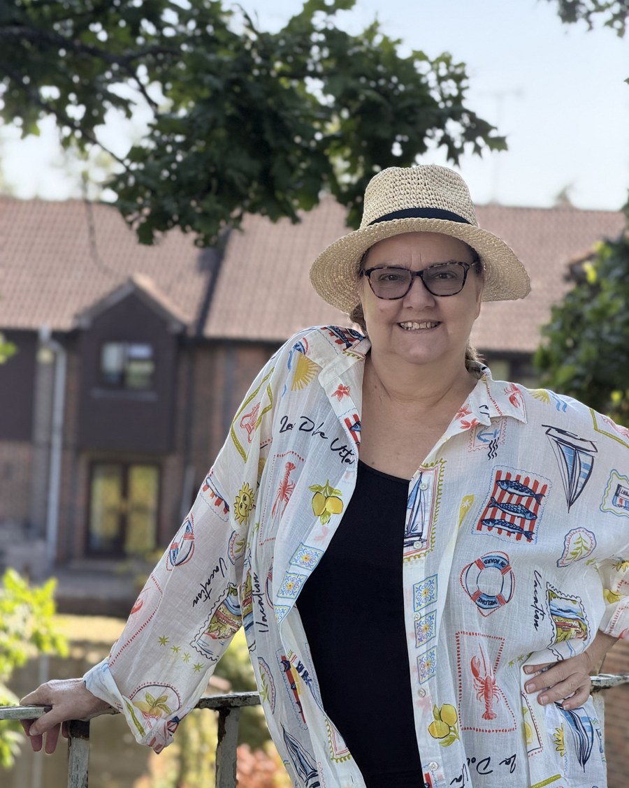 Jo-Dee Walmsley standing outdoors in a straw hat, smiling with her hand on her hip, leaning on a garden railing.